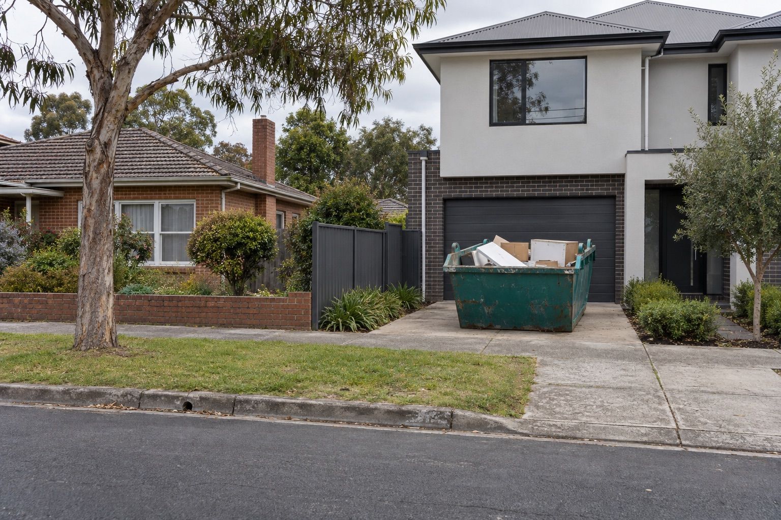 Skip bin hire, Bayswater: a teal skip of boxes and old panels on the driveway of a two-storey townhouse beside a brick house, on a flat street