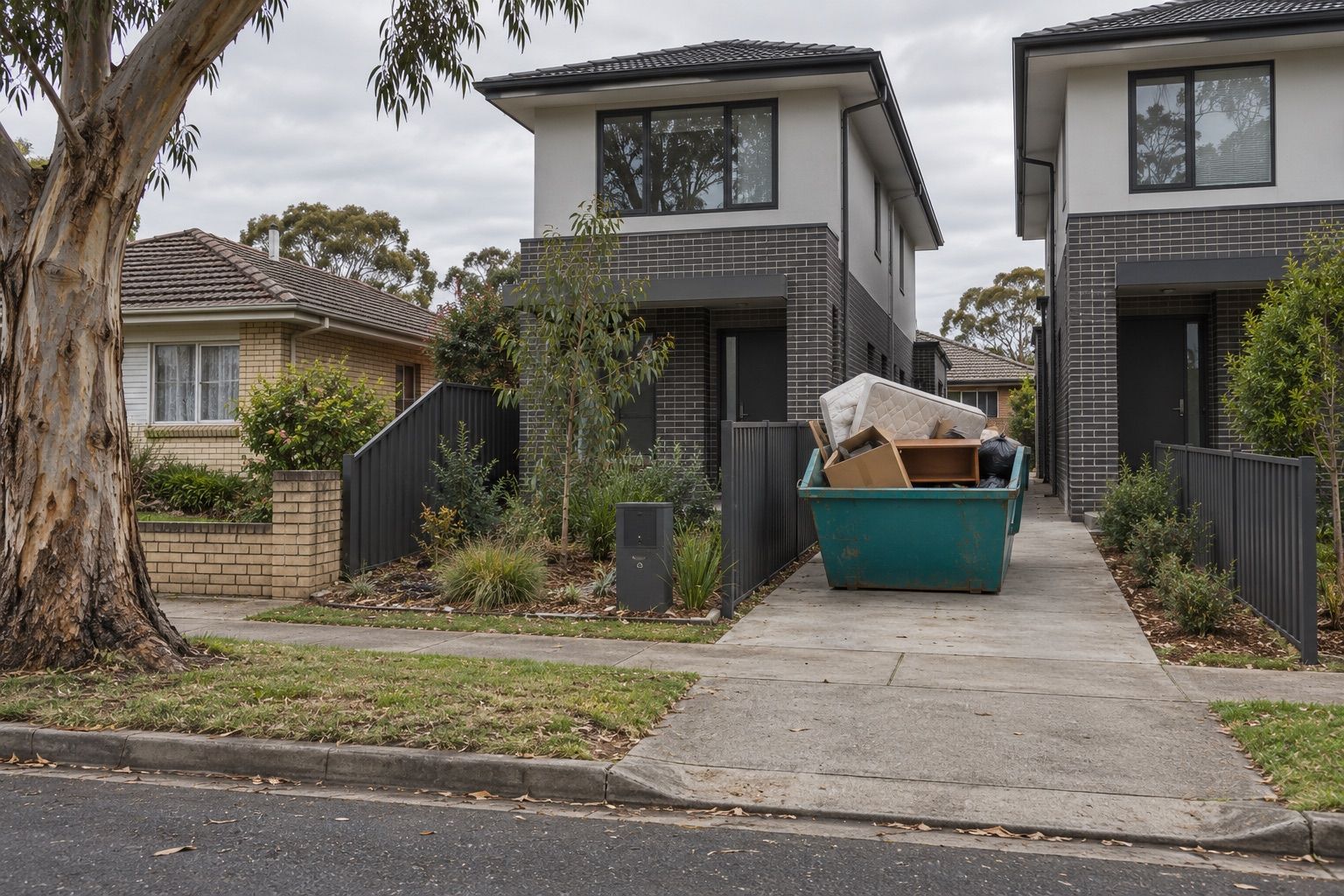Skip bin hire, Clayton: a small teal skip of boxes, a mattress and an old desk on the driveway of a new townhouse on a flat street