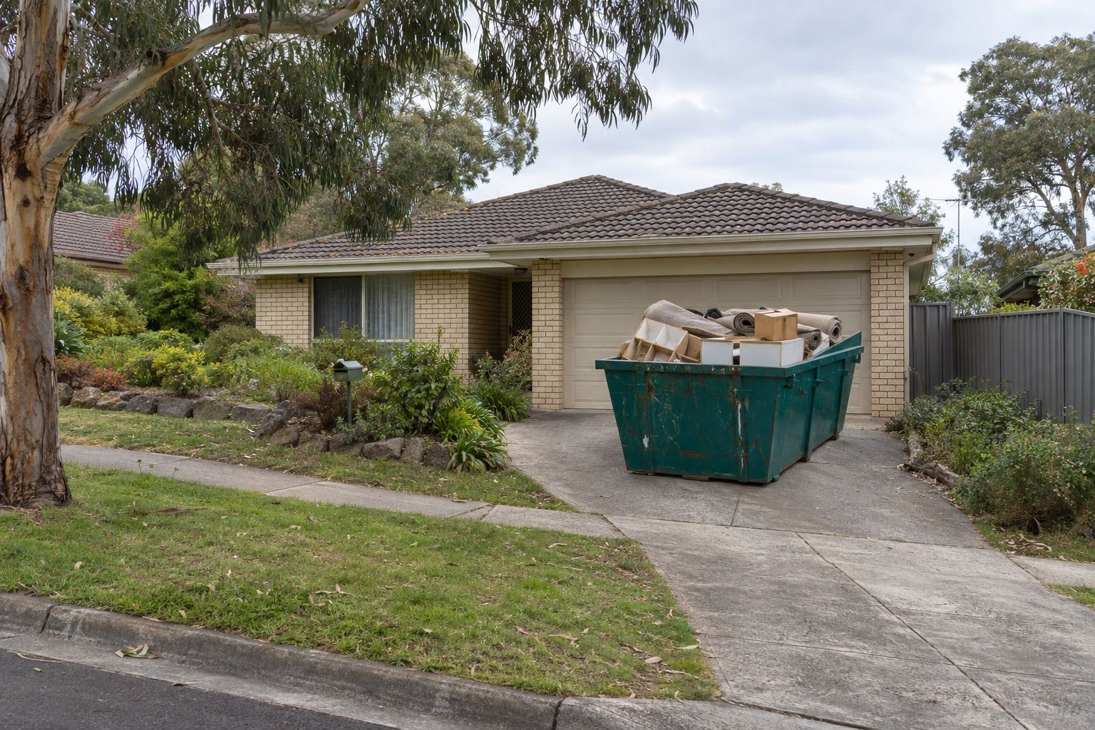 Skip bin hire, Mooroolbark: a teal skip of old carpet rolls and boxes in front of the garage of a brick-veneer family house