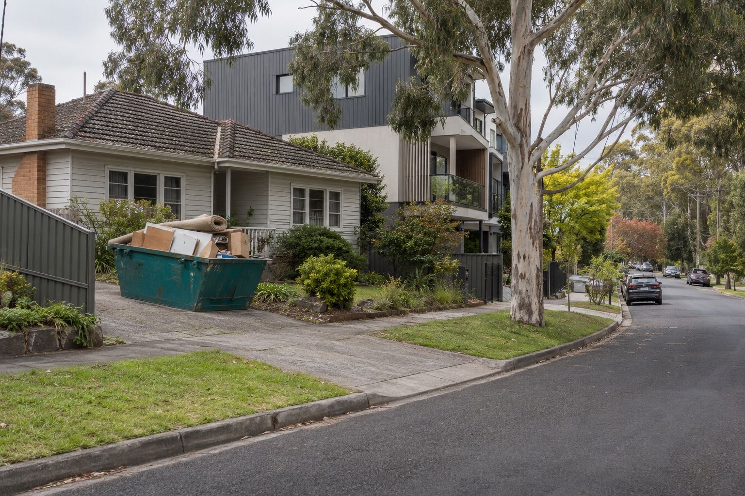 Skip bin hire, Ringwood: a small teal skip of flat-pack furniture, carpet and boxes on a weatherboard house's driveway beside new townhouses